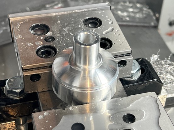 A part-finished aluminium mushroom shape with a hollow stalk to take a fine-pitch M12 thread.  This is help in a Gerardi milling vice and it has watery coolant beads splashed on the vice and part