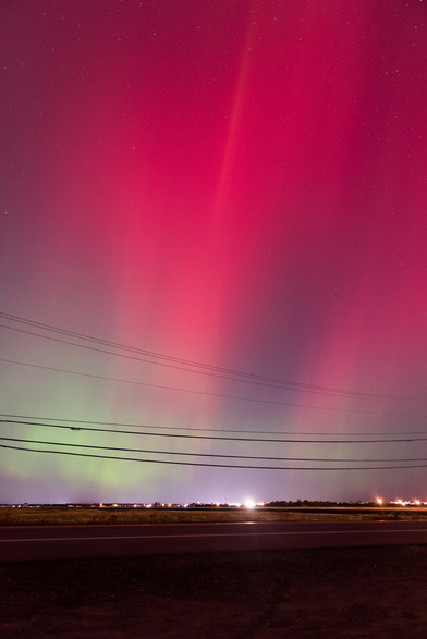 Photo of Oct 10th aurora borealis. Top half of the photo has pink/magenta tint with stars visible through it.  Below it, is a band of green tint but powerlines running left to right over it. Bottom quarter contains the ground - dirt in the immediate foreground, a 2 lane asphalt road beyond it, and march beyond that.  In the distance near the horizon are lights from various (1-2 story) buildings.
