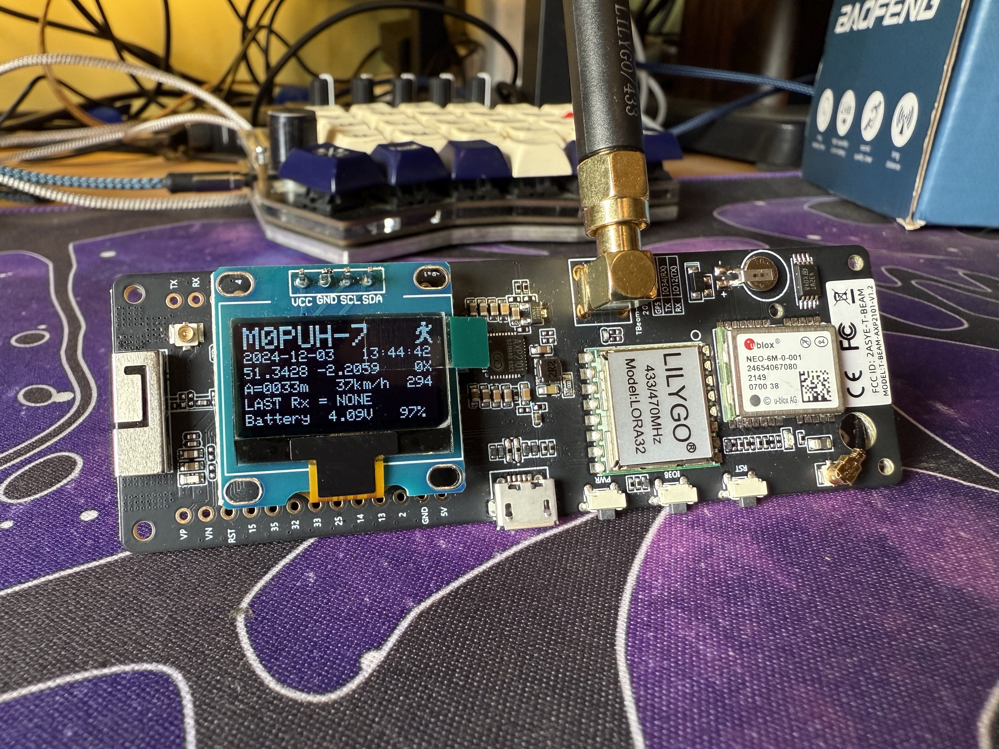 A small rectangular circuit board with a display on the left an antenna attached vertically to the middle and a couple of visible modules on the right (a GPS and one marked LILYGO). The display reads M0PUH-7 and had a date, time and battery voltage, among other details.