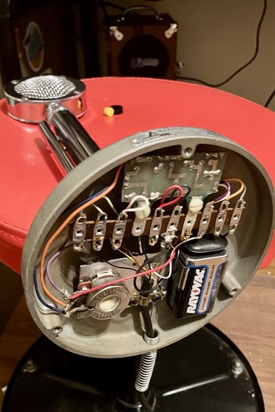 A chrome microphone lies on a red stool. The base of the microphone is open and shows a built-in amplifier circuit powered by a Rayovac 9v battery. 