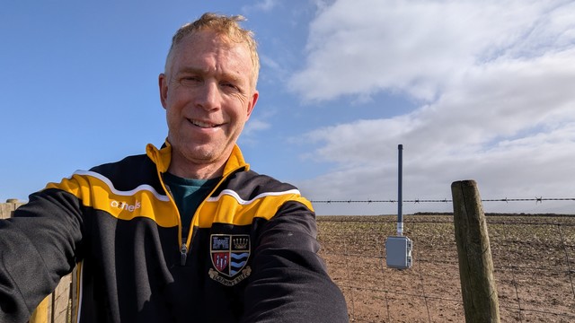 A slightly sweaty middle aged many wearing an Egremont rugby union top on. There is an APRS digipeater wedged into some fencing and this appears to be on farmland. The sweaty bloke is actually in a livestock pen on the top of a hill. The person is sweating because he had to cycle up a steep hill that had a gravel track and he's a bit unfit and has a cold (man-flu probably. In fact he should probably be tended to by a large medical team but it's mother's day)